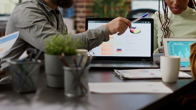 Diverse team of coworkers analyzing business charts and graphs on laptop and digital tablet, using networking software to plan presentation with diagrams. People working with research. Close up.