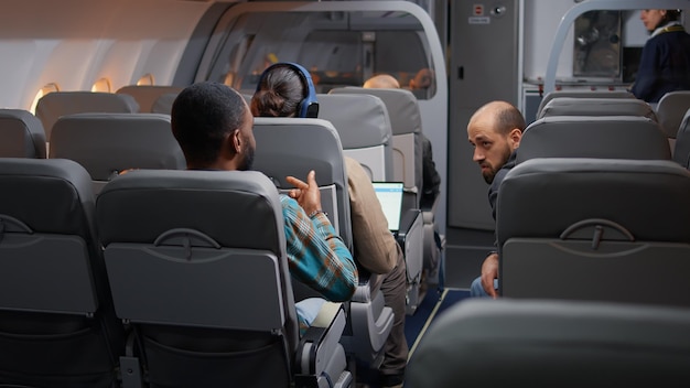 Разнообразные путешественники разговаривают в самолете перед взлетом, летят международной авиакомпанией, чтобы отправиться в отпуск. Путешествие на самолете с бортпроводником, коммерческие авиалинии.