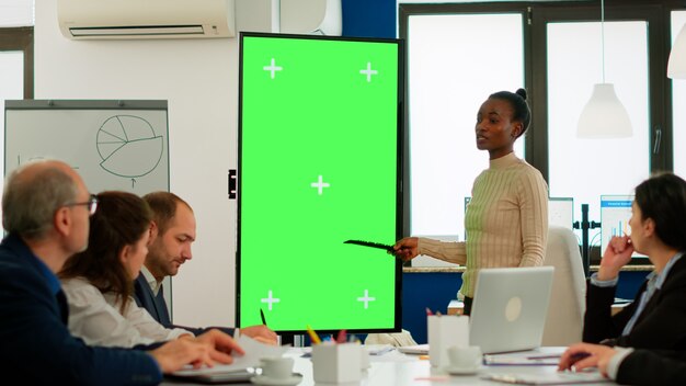ビジネスパートナーの前でグリーンスクリーンモニターと戦略を話し合うスタートアップオフィスに立っている多様な女性。多民族チームプロジェクトのクロマキーディスプレイモックアップデスクトップに説明するマネージャー