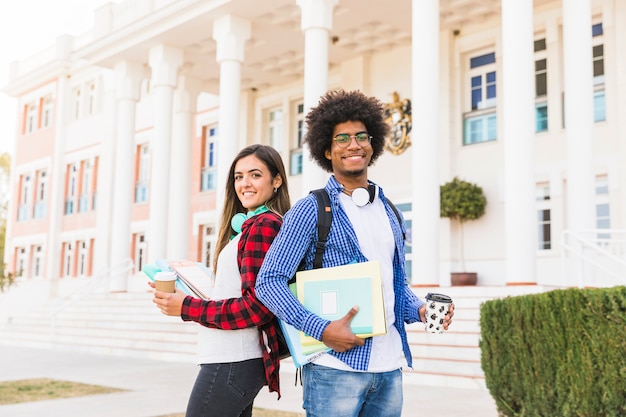 Diverse young male and female student holding books and takeaway coffee cup standing in front of building