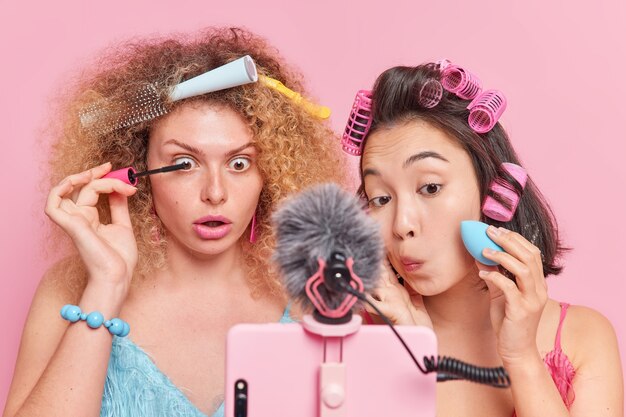 Diverse young women focused at smartphone webcam apply mascara and foundation gives make up tutorail to subscribers have own beauty blog present beauty cosmetics isolated on pink background.
