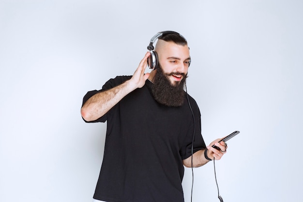 Dj with beard wearing headphones and setting music from his playlist at his smartphone.