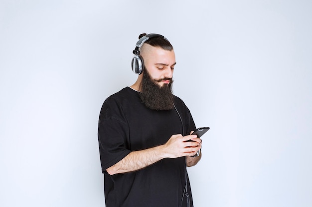Dj with beard wearing headphones and setting music from his playlist at his smartphone.