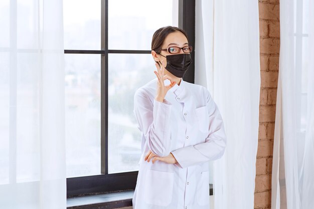 Doctor in black mask and eyeglasses standing by the window and showing enjoyment sign. 