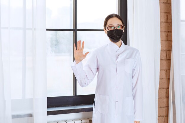 Doctor in black mask and eyeglasses standing by the window and stopping something. 