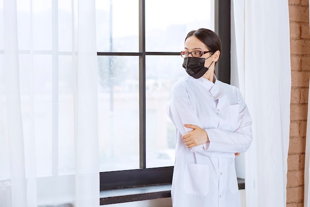 Doctor in black mask and eyeglasses standing by the window. 