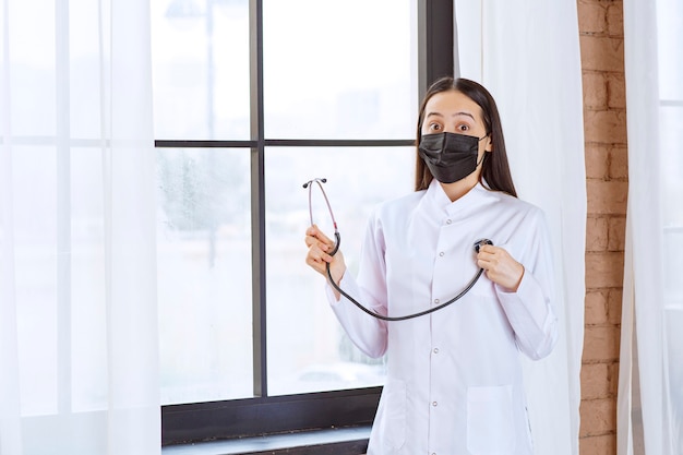 Доктор в черной маске со стетоскопом стоит у окна и проверяет сердцебиение.