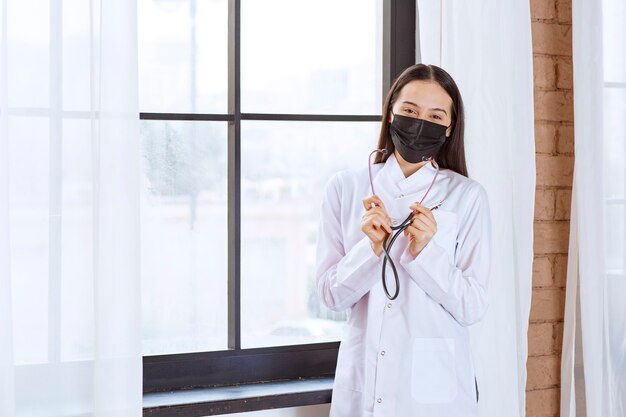 Doctor in black mask with a stethoscope standing by the window. 
