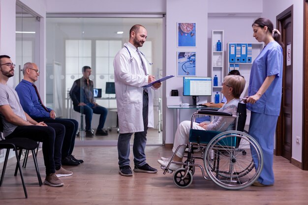 Doctor checking diagnosis of paralyzed disabled senior woman in wheelchair sitting in reception room...