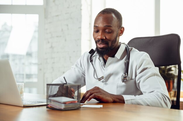 Doctor consulting for patient online, giving recommendation. African-american doctor during his work, explaining recipes for drug. Daily hard work for health and lives saving during epidemic.
