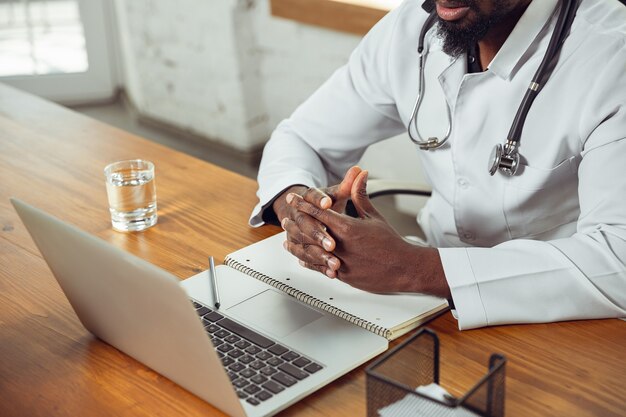 Doctor consulting for patient, working with laptop. African-american doctor during his work with patients, explaining recipes for drug. Daily hard work for health and lives saving during epidemic.