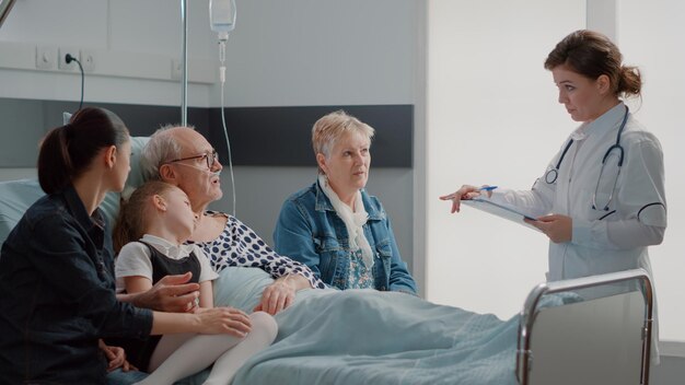 Doctor doing checkup visit with family visiting sick pensioner in hospital ward. Medic explaining diagnosis and treatment to relatives of senior patient, talking about healthcare.