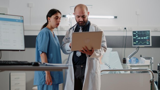 Doctor doing consultation with retired patient in bed. Medic with laptop talking to nurse about healthcare medicine before examining patient with nasal oxygen tube and heart rate monitor