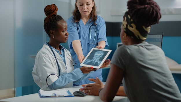 医療キャビネットの病気の患者にタブレットでX線スキャンを説明する医師。病気の女性に健康診断のためのデジタルデバイスでX線診断を示すメディック。