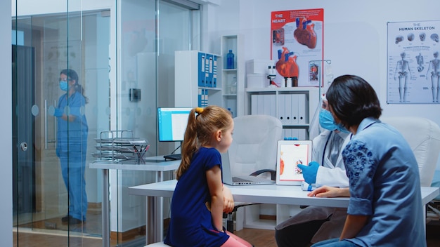 Doctor explining to child the functions of heart using tablet and wearing facial protection. Pediatrician specialist with gloves providing health care services consultations treatment during covid-19