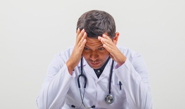 Doctor holding his head in his hands in white coat, stethoscope and looking thoughtful.