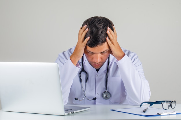 Free photo doctor holding his head in his hands in white coat, stethoscope and looking thoughtful ,