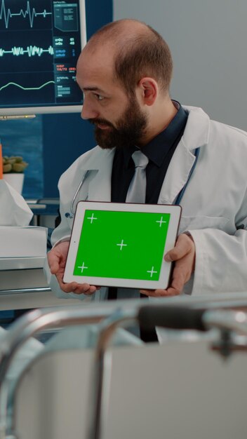 ベッドで病気の患者のためにデジタルタブレットで水平方向の緑色の画面を保持している医師。分離されたモックアップテンプレートと背景を持つデバイスのクロマキーを見ている医者と年配の女性