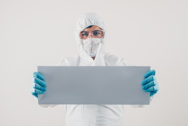 Доктор держа белый картон в медицинских перчатках и защитный костюм в светлой предпосылке. место для текстового коронавируса
