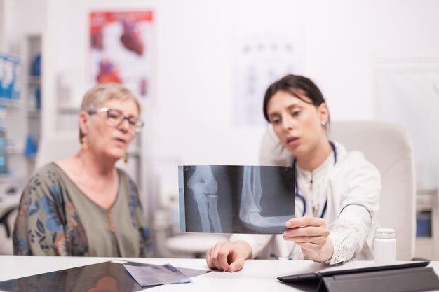 Doctor holding x-ray and talking with senior patient about knee injury in hospital office. Medic talking about therapy with mature woman.