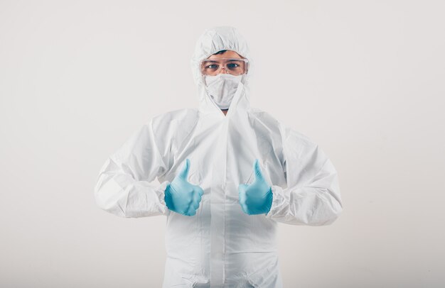 A doctor in medical gloves and protective suit showing thumbs up in light background . coronavirus