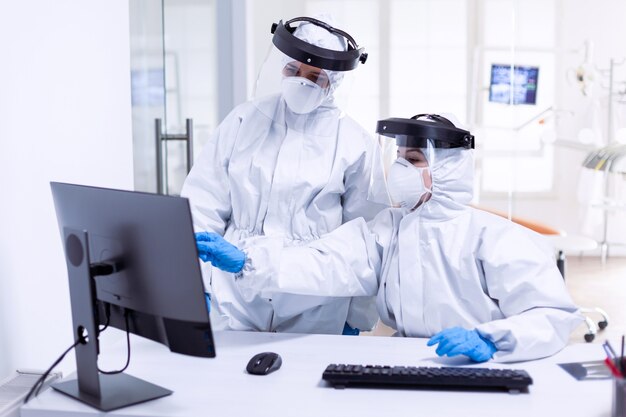 Doctor and nurse in ppe suit looking at monitor during global pandemic with covid-19. Medicine team wearing protection gear against coronavirus pandemic in dental reception as safety precaution.