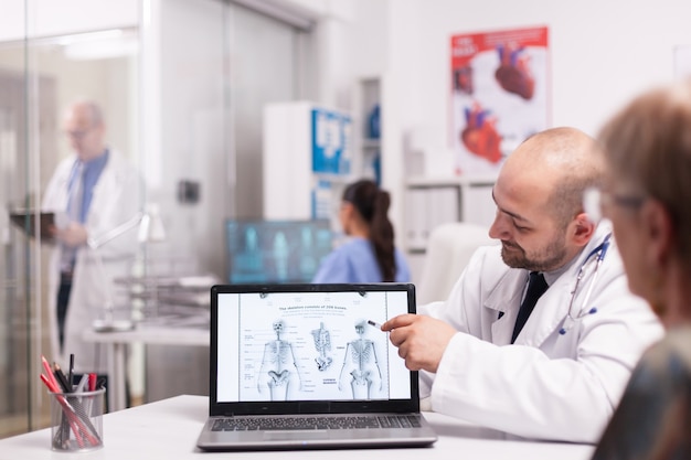 Doctor pointing at human skeleton on laptop in hospital office during consultation of old woman before surgery. Senior medic wearing white coat taking notes on clipboard in clinic corridor.