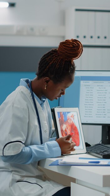 Doctor pointing at tablet with cardiovascular figure, explaining heart condition and blood flow to patient. Medic showing device screen with cardiac organ and cardiology diagnosis.