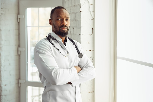 Доктор уверенно позирует в своем кабинете возле окна. Афро-американский врач во время работы с пациентами, объясняя рецепты лекарств. Ежедневная тяжелая работа для здоровья и спасения жизней во время эпидемии.