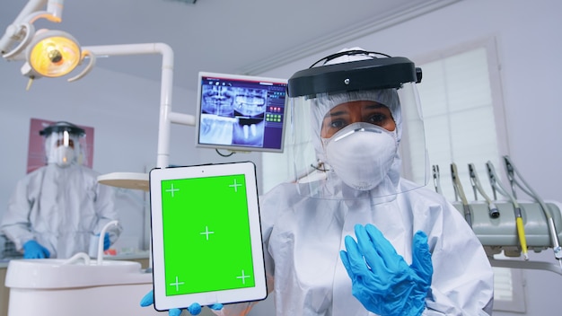 Doctor in ppe suit showing tablet with green screen, explaining dental radiography and diagnosis for teeth infection. Stomatology specialist in coverall pointing at mockup, copy space, chroma display