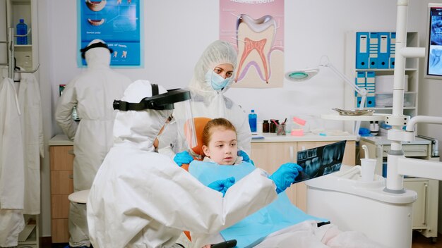 Doctor in protective suit discussing about x-ray of tooth with mother of child patient explaining treatment using tablet in covisd-19 pandemic. Medical team wearing face shield, coverall, mask, gloves