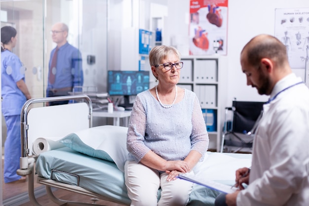 Doctor reading diagnosis for sick senior woman sitting on hospital bed in examination room