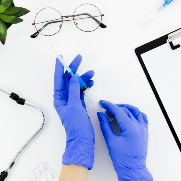 Рука доктора нося голубые перчатки держа шприц на белом столе с стетоскопом; очки; таблетки и буфер обмена