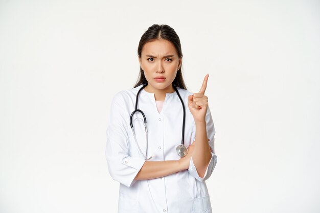 의사는 환자에게 화난 화난 여성 아시아 의사가 손가락을 흔들고 눈을 찡그리며 불만스럽게 꾸짖는 모습을 보여줍니다.