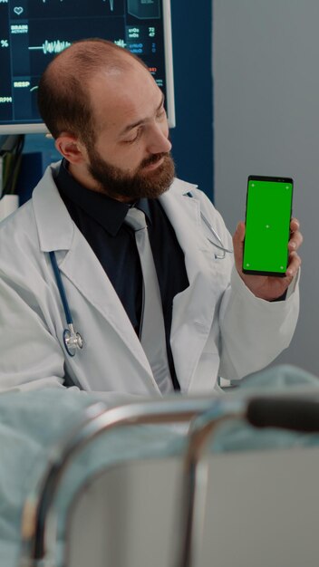 看護施設で電話で緑色の画面を見ている医師と病気の患者。分離されたテンプレートとモックアップの背景のクロマキーでスマートフォンを垂直に保持している医療専門家