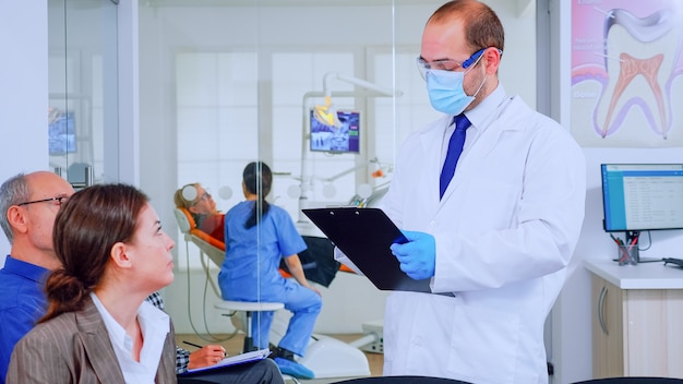 의사는 구강 클리닉의 대기실에서 의자에 앉아 있는 환자의 치과 문제에 대해 클립보드에 메모합니다. 백그라운드에서 검사를 위해 노파를 준비하는 조수