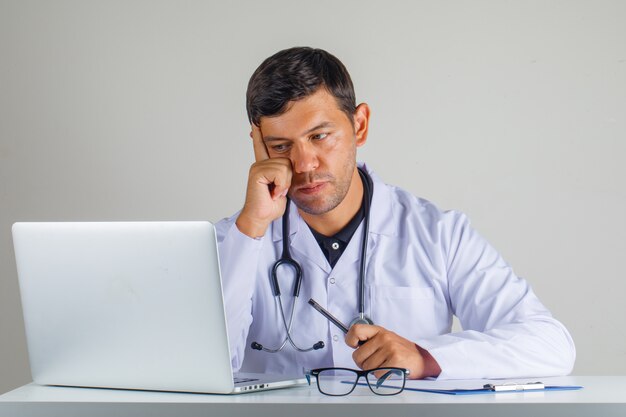 Doctor in white coat, stethoscope sitting and looking at laptop and looking careful