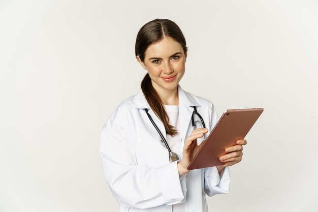 Врач в белом халате с помощью цифрового планшета читает медицинские данные на гаджете, работающем в больнице, стоящей...