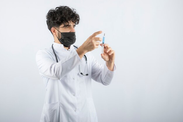 Doctor in white coat with stethoscope holding and touching a syringe isolated on white. 