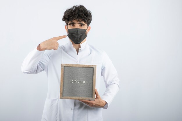 Doctor in white coat with stethoscope pointing at mask isolated on white. 