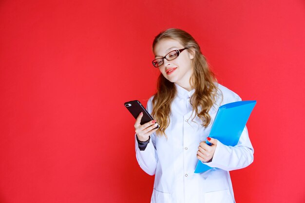 Doctor with a blue folder and eyeglasses checking her telephone.