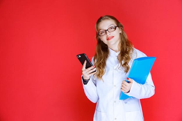 Doctor with a blue folder and eyeglasses checking her telephone.