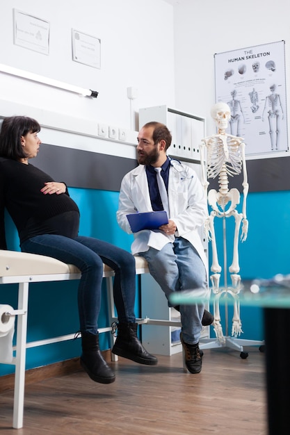 Doctor with clipboard consulting pregnant woman at exam appointment in office. Expectant patient discussing about childbirth and pregnancy with physician, receiving support at checkup visit.