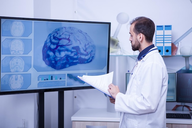 大きな​モニター​で​脳​を​見ている​彼​の​手​に​クリップ​ボード​を​持っている​医者​。​自信​の​ある​医者​。​制服​を​着た​医者​。