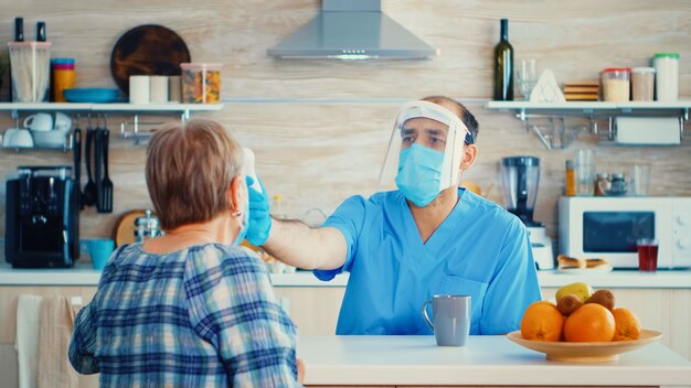 Doctor with face mask checking senior woman temperature using gun thermometer during home visit. Social worker visiting vulnerable persons for disease spreading prevention during COVID-19 campaign