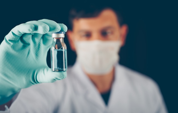 Doctor with gloves and mask holding vial in his hand close-up
