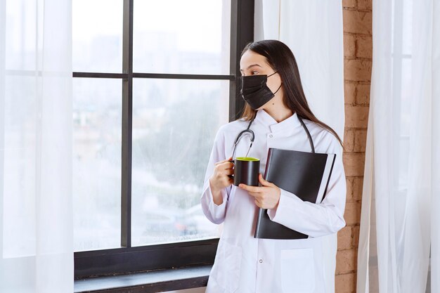 Doctor with stethoscope and black mask holding a black cup of drink and a black folder and looking through the window. 