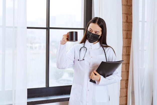 Doctor with stethoscope and black mask holding a black cup of drink and a black folder. 