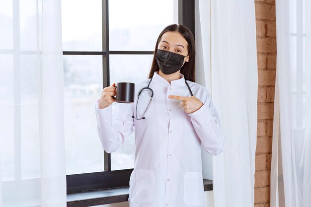 Doctor with stethoscope and black mask holding a black cup of drink at the break. 