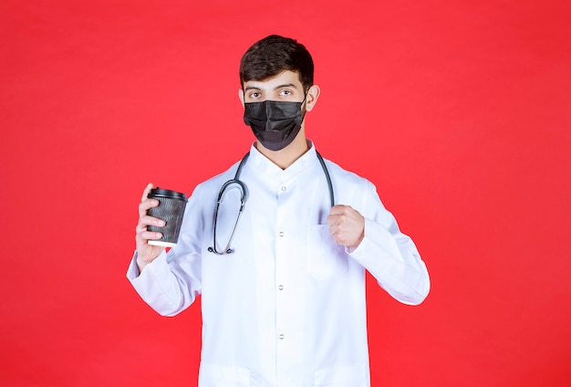 Doctor with stethoscope in black mask holding a black takeaway coffee cup and showing his fist. 